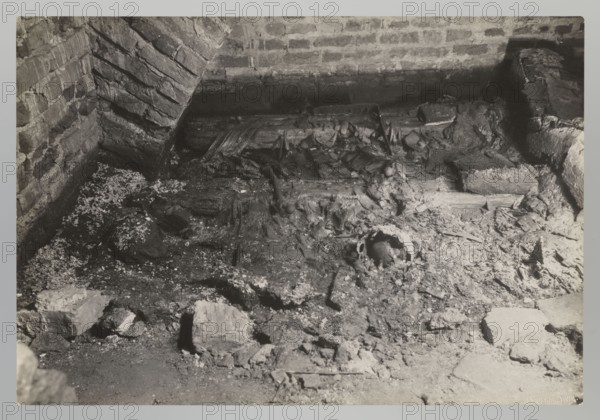 Vilnius. Cathedral — interior of the royal crypt, left side; Bulhak, Jan 1876–1950; Vilnius Lithuania; Archcathedral Basilica of St. Stanislaus and St. Ladislaus; crypts; archaeological excavations following the flood in Vilnius in 1931; 1931; photograph natural; 11.5 × 17.0 cm, The image shows a rough interior corner with brick walls and an arched brick support, a floor area covered in rubble, broken wood fragments, and scattered debris. A few larger stone or brick chunks lie amid finer dusty material and ash-like particles. Textural details include cracked and flaking surfaces on the bricks, splintered timber, and crumbly masonry. Visible colors are shades of gray, black, and off-white.