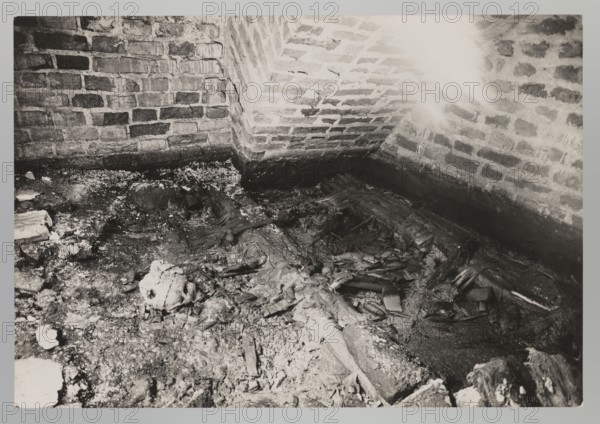 Vilnius. Cathedral — interior of the royal crypt. Photograph by Jan Bulhak 1876–1950, Vilnius, Archcathedral Basilica of St. Stanislaus and St. Ladislaus, crypts, archaeological excavations; Alexander I Jagiellon King of Poland; 1461–1506 mentioned. Flood in Vilnius in 1931; 1931. Photograph from nature. Image size: height 11.5 cm, width 16.7 cm., The image shows a corner of a room with brick walls and a dirt floor; the bricks are various shades of gray and dark gray, and there are lighter gray patches where mortar shows. Scattered across the floor are decomposed organic materials and debris in tones of dark gray, medium gray, and pale gray, with a discernible skull-like shape among the rubble. The scene includes fragments of wood and rubble in darker and lighter gray hues, areas of crumbling texture, and a lighter glare on the upper right brick wall. Overall the visible colors are shades of gray, black, and white.