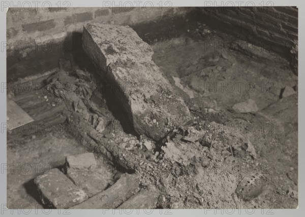 Vilnius. Cathedral — interior of the royal crypt; photograph from nature by Jan Bulhak 1876–1950; Archcathedral Basilica of St. Stanislaus and St. Ladislaus, crypts; archaeological excavations; related to Barbara Radziwillówna Queen of Poland, 1520–1551; flood in Vilnius, 1931; photograph, 1931; dimensions: height 11.5 cm, width 15.5 cm., The image shows a close-up of a rough, dusty scene with a large broken stone or concrete slab leaning at an angle over a crumbling base surrounded by debris and fragments; a group of smaller bricks and rubble lie in the foreground, and a textured wall with horizontal lines appears in the background. The visible colors are shades of gray, black, and off-white.