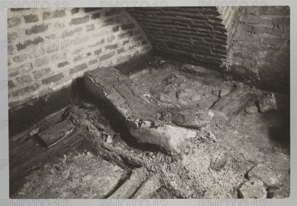 Vilnius. Cathedral — interior of the royal crypt, Bulhak, Jan 1876–1950; Archcathedral Basilica of St. Stanislaus and St. Ladislaus Vilnius; crypts; archaeological excavations; Barbara Radziwillówna Queen of Poland; 1520–1551; flood in Vilnius in 1931; 1931; Vilnius; photograph from nature; photograph; height 11.5 cm; width 16.7 cm, The image shows an indoor corner with exposed brick walls meeting at an angle and a floor covered with dirt, rubble, and several pieces of broken wood and stone; a few larger, rough wooden beams lie across the floor, one of which is cracked and partially embedded in a mound of debris, and there are a few scattered small stones and fragments around it. The visible colors are shades of gray, black, and off-white.