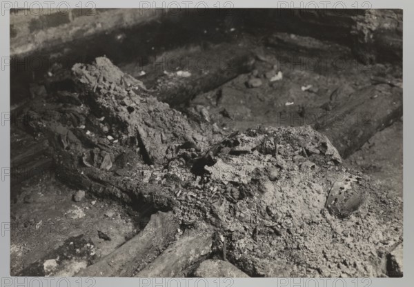 Vilnius. Cathedral — interior of the royal crypt. Photograph from nature by Jan Bulhak 1876–1950, 1931. Depicts the crypts and archaeological excavations, including association with Barbara Radziwillówna Queen of Poland; 1520–1551. Height 12.0 cm; width 16.5 cm., The image shows a close-up of an irregular, rough textured surface with a group of decomposed, layered materials and debris spread across a ground plane; the scene is rendered in shades of black, white, and gray, with darker areas in the background and lighter, ashy patches and granular accumulations in the foreground; there are elongated, cylindrical forms partially embedded in the debris and a rounded object with a mottled surface visible among the lighter material; overall the composition features cracked, crumbly textures, scattered fragments, and contrasting tonal areas of dark gray, light gray, and near-white.