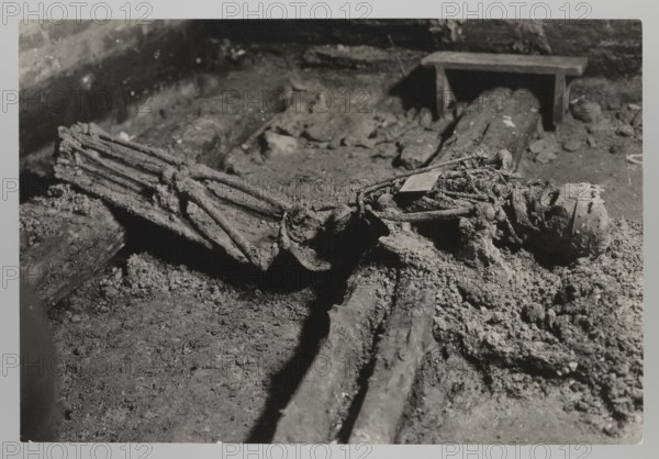 Vilnius. Cathedral — interior of the royal crypt. Photograph from nature by Jan Bulhak 1876–1950, 1931. Depicts the crypts and archaeological excavations, including association with Barbara Radziwillówna Queen of Poland; 1520–1551. Height 11.5 cm, width 17.0 cm., The image shows a close-up scene in muted grayscale tones of a textured ground surface with a group of decomposed organic materials and wooden beams lying across it; several frayed rope-like strands and fibrous bundles rest atop and around the beams, creating irregular clumps and rough granular debris. A cylindrical object with an open end and patterned edge lies partially embedded in the debris near the right side, while a flat rectangular board is visible in the background supported by short legs, casting a shadow. The overall palette is shades of black, white, and gray with varied contrasts between the lighter granular deposits and the darker shadows and wood surfaces.