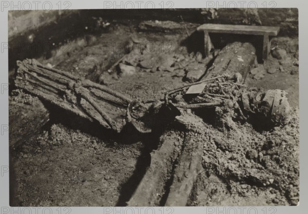 Vilnius. Cathedral — interior of the royal crypt; photograph from nature by Jan Bulhak 1876–1950, 1931. Archcathedral Basilica of St. Stanislaus and St. Ladislaus Vilnius, crypts; archaeological excavations; burial of Barbara Radziwillówna Queen of Poland; 1520–1551; flood in Vilnius in 1931. Photograph, 11.5 × 17.0 cm., The image shows a close-up scene dominated by textured, rough surfaces in shades of black, gray, and off-white, featuring an arrangement of elongated, cylindrical objects lying on a dirt-covered ground with granular debris scattered around. A group of rope-like strands and bands are visible, appearing encrusted and tangled, alongside a few rigid, plank-like pieces. In the background there is a simple bench-like structure casting a shadow, and the overall lighting creates high contrast with deep shadows and bright highlights across the soil and objects.