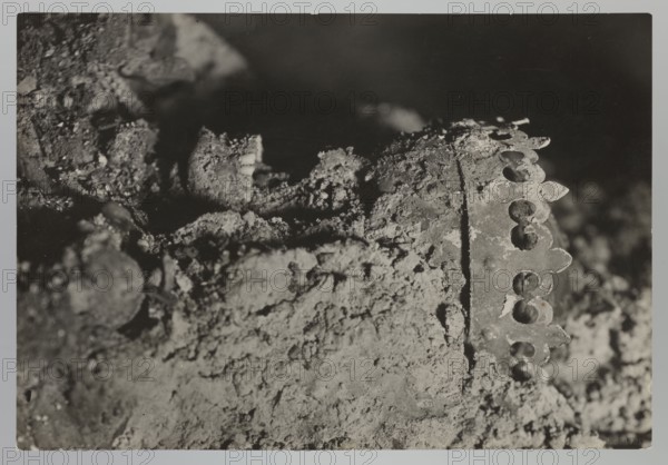 Vilnius. Cathedral — interior of the royal crypt. Photograph from nature by Jan Bulhak 1876–1950, 1931. Subject: Archcathedral Basilica of St. Stanislaus and St. Ladislaus Vilnius, crypts; archaeological excavations; Barbara Radziwillówna Queen of Poland; 1520–1551. Flood in Vilnius 1931. Place of origin: Vilnius Lithuania. Medium: photograph. Dimensions: height 11.5 cm, width 16.5 cm., A black-and-white photograph showing a close-up of a rough, textured surface covered in crumbly material and granular deposits, with a metallic decorative edge partially embedded in the debris on the right side; the metallic piece has a scalloped pattern with oval cutouts and visible rivets, while the rest of the scene includes irregular clumps and fragments creating deep shadows and highlights in shades of black, gray, and white.