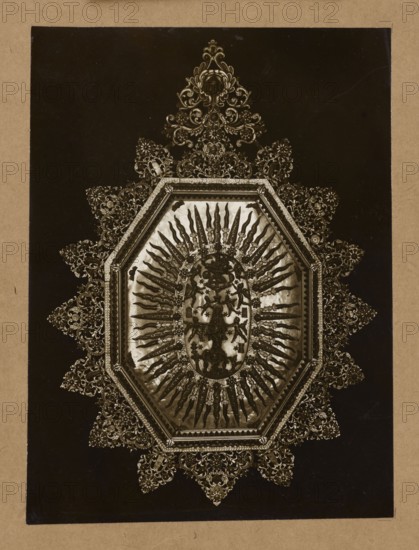 Coral altar-reliquary with a depiction of the Assumption of the Virgin Mary, 17th century — general view; materials: coral; function: altar-reliquary; subject/iconography: Assumption of the Virgin Mary, mandorla, angels, plant tendril; style/genre: jewelry, reliquary, altars;, physical description of photograph: photographic print from nature, entire surface re-glued to cardboard; photo dimensions: print height 20.5 cm, width 15.2 cm; cardboard height 34.8 cm, width 25.0 cm; unknown creator/photographer., The image shows an ornate octagonal frame with intricate filigree extending outward in a starburst pattern against a dark background; the central area contains a decorative oval with radiating elements that resemble elongated rays or blades. Colors visible include shades of brown, sepia, gold, black, and beige, with the metallic decorative details appearing golden against darker brown and black areas, and a beige border surrounding the dark central panel.