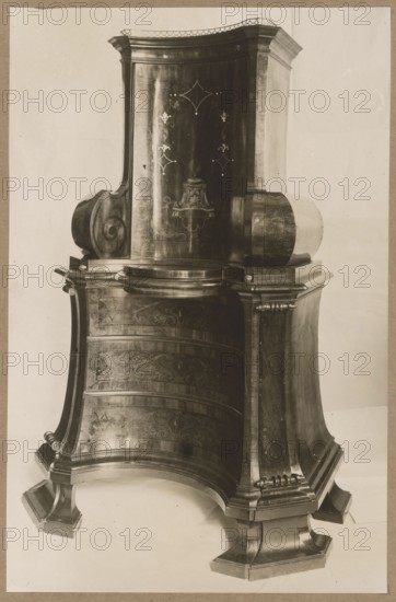 Two-part sacristy cabinet with a semicircular concave front, 2nd quarter of the 18th century; Baroque furniture with intarsia, church cabinet; photographic reproduction, photographic print height 21.8 cm, width 14.1 cm; cardboard height 34.8 cm, width 24.8 cm; photographer possibly Józef Kosciesza Jaworski fl. ca. 1911–ca. 1929., The image shows a large ornate wooden piece of furniture with a curved, sculpted form and decorative inlay patterns. The object has a tall central section with a rounded top and carved side scrolls, a broad base with recessed panels and curved legs, and a stepped plinth. Visible colors include shades of brown, beige, and dark brown tones, with highlights creating lighter beige and off-white reflections on the polished wood surfaces. A plain light background contrasts with the darker furniture.