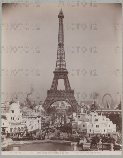 Exposition universelle de 1900 - Tour Eiffel et Champ de Mars Eiffel Tower and Champ de Mars, Jean Gilletta 1856–1933, photograph, 1900. Silver gelatin/photographic print glued across entire surface onto cardboard backing; print 29.1 × 23.0 cm, cardboard 42.5 × 33.0 cm. Subject: Eiffel Tower, Champ de Mars, Paris; World Exhibition Paris, 1900. Style: photography from nature; architecture French/European., The image shows a large iron lattice tower rising centrally against a pale sky, with a wide plaza and a procession of people and structures leading toward its base. In the foreground there are several pale buildings with domed and arched roofs, and a body of water with stone balustrades and sculpted figures near the edge. The plaza is filled with a dense group of people, pathways, and a mix of pavilions and tents with varied roof shapes. Behind and around the tower are additional architectural features, including domes, spires, and a large circular wheel structure, and faint plumes of...