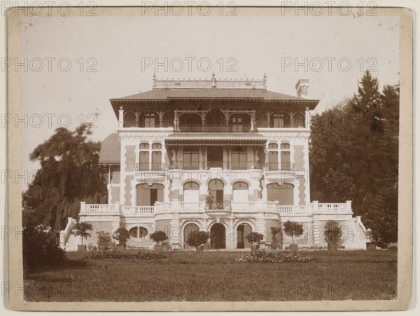 Ignacy Jan Paderewski's villa in Riond-Bosson in Switzerland — general view from the garden, unknown photographer; subject: Paderewski, Ignacy Jan 1860–1941; location: Riond-Bosson, Morges, Switzerland; themes: villas, architecture, European architecture, gardens, Swiss architecture, palaces, foreign architecture;, date: ca. 1900; medium/technique: photography from nature, photographic print glued across the entire surface to cardboard; format: print height 16.7 cm, width 22 cm; support height 17.9 cm, width 23.8 cm., A large multi-story building with ornate balconies and arched windows sits centered in the image, flanked by trees on both sides. The façade shows decorative railings, columned porches, and curved bay window elements, with a wide terrace and balustrade at the main level and staircases descending at the sides. In front of the building is a lawn with a flowerbed and several potted and planted small trees or shrubs along the terrace. The scene is rendered in sepia tones with...