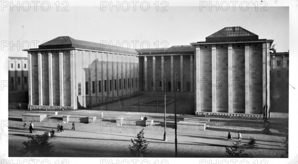 View of the central pavilions of the National Museum at Aleja 3 Maja, Warsaw; photograph by Photo-Plat photographic studio, 1927–1939. Shows museum buildings, street lamps, surrounding architecture on Aleja 3 Maja/Aleje Jerozolimskie, Warsaw; Polish architecture; dated 1938. Photography nature photography, 10.2 cm × 17.9 cm., A large neoclassical building with tall vertical columns and flat roofs sits behind an open paved plaza; the building and plaza appear in shades of black, white, and gray, with shadows creating darker gray areas. In the foreground the plaza shows a few low rectangular stone benches and stepped platforms, and a group of people walking and sitting; their clothing appears as dark and light gray silhouettes. Tall slender street lamps rise from the plaza casting long diagonal shadows. A few small trees with dark gray foliage are visible near the bottom edge. The sky above is a pale gray expanse.