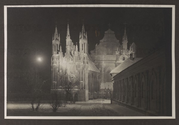 Vilnius Illuminated. Churches of St. Anne and the Bernardines; Edmund and Boleslaw Zdanowski, photographers; Vilnius Lithuania, Bernardine Church, St. Anne's Church; before 1939; photograph, photographic print: height 8.5 cm, width 13.5 cm; album page: height 17.0 cm, width 24.5 cm, A nighttime scene showing a large gothic-style building with tall spires and ornate architectural details illuminated against a dark sky, a smaller building with a sloped roof and a row of arched openings extending along the right side, a snow-covered foreground with a few leafless trees and low shrubs, a bright light source in the sky casting light across the scene, and visible tones of black, dark gray, light gray, and white.