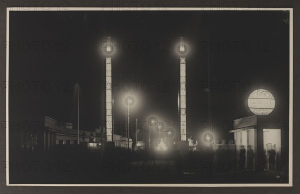 Vilnius Illuminated. Northern Fair, photographed by Edmund and Boleslawa Zdanowscy, Vilnius Lithuania, Northern Fair Vilnius, before 1939, photograph, photographic print: height 8.5 cm, width 13.5 cm, album page: height 17.0 cm, width 24.5 cm, The image shows a night scene with a dark sky and illuminated vertical structures emitting circular glows at their tops; there are a few tall illuminated pillars in the foreground and several smaller glowing streetlights receding into the distance, creating rings of light. A group of low buildings with flat roofs runs along the lower part of the scene, and a rectangular entrance structure with a bright interior light stands to the right where a few blurred human figures are visible. The visible colors are black, white, and shades of gray.