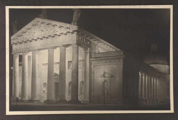 Vilnius Illuminated. Cathedral, facade and southern elevation, photographed by Edmund and Boleslawa Zdanowscy; Vilnius Lithuania; cathedral Vilnius; classicism architect; before 1939; photograph, photographic print: height 8.5 cm, width 13.5 cm; album page: height 17.0 cm, width 24.5 cm, A nighttime scene of a classical building facade with a row of large, round columns supporting a triangular pediment; the facade and columns are lit in pale tones while the background is dark, creating strong contrasts of light and shadow. The pediment and entablature show carved reliefs and decorative details. Between the columns are recessed niches each containing sculptural figures, and there is a taller rectangular section to the right with a recessed statue in a framed niche and a slender street lamp in front. To the far right a colonnaded extension with several columns leads toward a domed structure partially visible behind it. The image shows shades of black, gray, cream, and sepia.