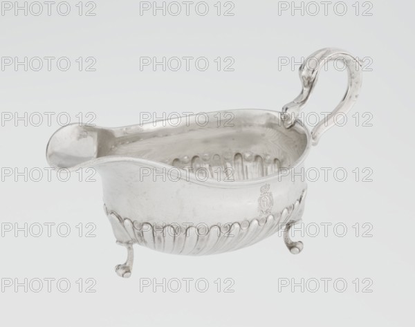 Sauce boat by Charles Stuart Harris fl. 1852–1897, made by Harris, CS & Sons London; goldsmiths, fl. 1817–1933. Oval sauce boat supported on three volute legs with an S-shaped handle and a wide rim on the axis. Bottom decorated with alternating convex and concave clasps. Side engraved with the Leszczyc Radolinski coat of arms; bottom inscribed II RAD MAJ I 747. Dates: 1892/1893. Great Britain — England, London. Vessel. Height 10 cm; width 22.5 cm; weight 167.5 g., A single polished metal sauce boat with a reflective surface sits against a pale background, showing a smooth curved pouring spout at one end and an ornate scroll-shaped handle at the other; the body has a scalloped lower edge with a row of rounded vertical ridges and sits on a group of small curved feet, and the visible colors are silver tones with subtle gray reflections and a very light neutral backdrop.
