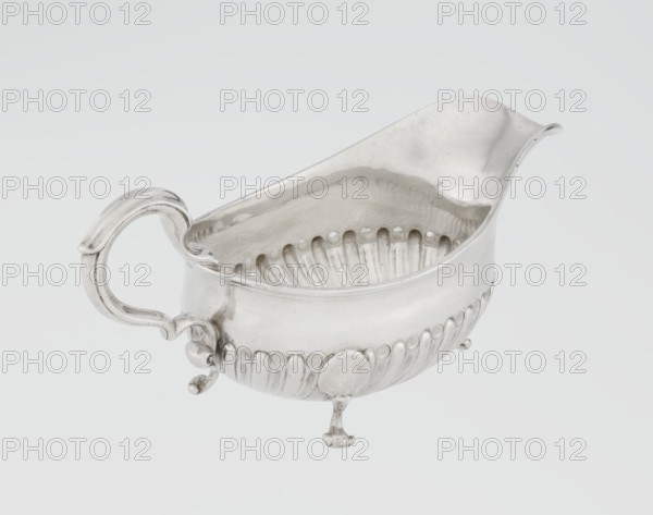 Sauce boat by Charles Stuart Harris fl. 1852–1897 and Harris, CS & Sons London; goldsmith's firm; fl. 1817–1933. Oval sauce boat supported on three volute legs with an S-shaped handle and a wide rim on the axis. Bottom decorated with alternating convex and concave clasps. Side engraved with the Leszczyc Radolinski family coat of arms; inscription on the bottom: II RAD MAJ I 747, 1892/1893. Made in London, England. Vessel; height 10 cm, width 22.5 cm, weight 167.5 g., A silver-colored metal sauce boat with a curved pouring spout and an ornate handle sits against a pale gray background; the boat has a rounded body with embossed vertical fluting along the lower exterior and a scalloped interior rim, resting on three small decorative feet, and the surface shows reflective highlights and subtle shadows in shades of silver, gray, and white.