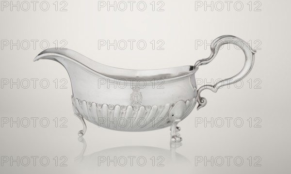 Gravy boat, maker: Charles Stuart Harris fl. 1852–1897 and firm Harris, CS & Sons London; goldsmith's firm; fl. 1817–1933. Oval gravy boat supported on three volute legs with S-shaped handle; wide rim on the axis. Bottom decorated with alternating convex and concave clasps. Side engraved with the Leszczyc Radolinski family coat of arms; bottom inscribed II RAD MAJ I 748. Dated 1892/1893. Silver vessel, London, England; height 10 cm, width 22.5 cm, weight 167.5 g., A silver-colored metal sauce boat with a smooth, curved rim and pointed pouring spout sits on a pale, neutral background; the body has a rounded, fluted lower section and an embossed decorative band near the top, supported by a few ornate feet that create a subtle reflection on the surface beneath, and a large, curving handle with flowing, sculpted lines extends from one side.