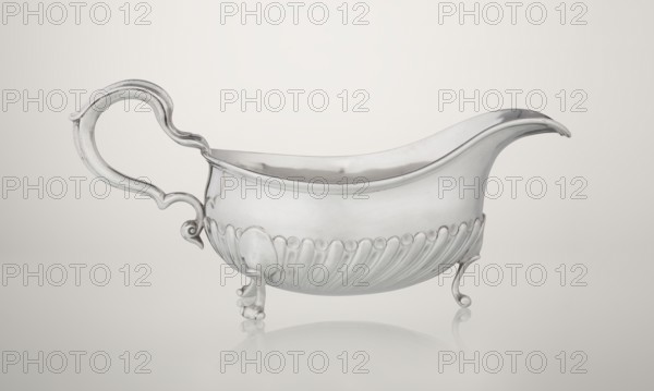 Gravy boat. Maker: Charles Stuart Harris fl. 1852–1897; firm: Harris, CS & Sons London; goldsmith's firm; fl. 1817–1933. Oval gravy boat supported on three volute legs, S-shaped handle, wide rim on the axis. Bottom decorated with alternating convex and concave clasps. Side engraved with the Leszczyc Radolinski family coat of arms; bottom inscribed II RAD MAJ I 748. Date: 1892/1893. Place: London, England, Great Britain. Vessel; height 10 cm, width 22.5 cm, weight 167.5 g., A silver-colored metal sauceboat with a curved spout and an ornate scrolling handle sits on a reflective pale background; the body has a smooth upper surface and a lower section decorated with a row of rounded bead-like shapes above fluted drapery, and it stands on a few small ornate feet that are also silver in color, with subtle highlights and shadows in grey and white across the vessel.