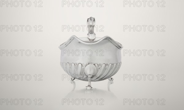 Gravy boat, maker: Charles Stuart Harris fl. 1852–1897 and Harris, C.S. & Sons London; goldsmith's firm, fl. 1817–1933. Oval gravy boat supported on three volute legs with an S-shaped handle; wide rim on the axis. Bottom decorated with alternating convex and concave clasps. Side engraved with the Leszczyc Radolinski family coat of arms; bottom inscribed II RAD MAJ I 748. Date: 1892/1893. Great Britain — England, London. Vessel; height 10 cm, width 22.5 cm, weight 167.5 g., A reflective silver bowl with a scalloped rim and ornate handle sits on a pale smooth surface, supported by several small curved legs; the bowl shows decorative vertical grooves on its lower half and has a soft reflection beneath it, with colors mainly silver, white, and light gray, and subtle darker gray shadows.