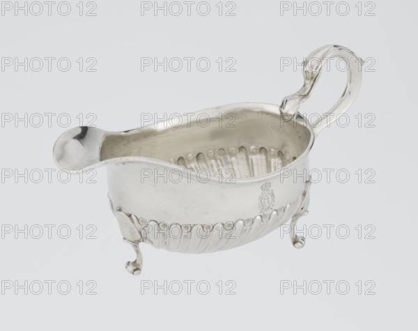 Gravy boat, maker: Charles Stuart Harris fl. 1852–1897 and firm Harris, CS & Sons London; goldsmith's firm; fl. 1817–1933. Oval gravy boat supported on three volute legs, S-shaped handle; wide rim on the axis. Bottom decorated with alternately convex and concave clasps. On the side, engraved coat of arms of the Leszczyc Radolinski family; on the bottom, inscription II RAD MAJ I 748. Dated 1892/1893. London, England, Great Britain. Vessel: height 10 cm, width 22.5 cm, weight 167.5 g., A polished silver sauce boat with a curved spout and an ornate looping handle sits against a plain pale gray background, showing a reflective interior and rounded rim; the body features a scalloped lower edge with a row of raised oval motifs and vertical fluted panels, and it stands on a group of three curled cabriole legs with scroll feet.
