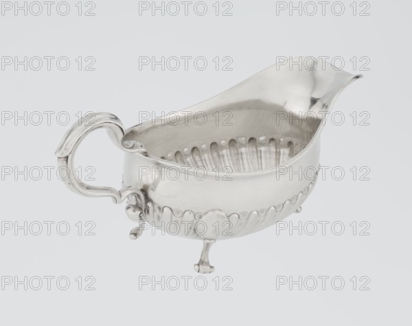 Oval gravy boat, supported on three volute legs, S-shaped handle; wide rim on the axis. Bottom decorated with alternately convex and concave clasps. On the side, engraved coat of arms of the Leszczyc Radolinski family; on the bottom, inscription II RAD MAJ I 748. Maker: Charles Stuart Harris fl. 1852–1897 and Harris, CS & Sons London; goldsmiths. Date: 1892–1893. Material/format: silver vessel. Dimensions: height 10 cm, width 22.5 cm, weight 167.5 g. Place: London, England, Great Britain., A silver-colored metal gravy boat with a curved spout and an ornate handle sits against a light gray background; the interior and exterior surfaces are reflective with polished highlights and soft shadows, the exterior features a scalloped band of raised oval decorations and fluted vertical grooves along the lower half, and the piece rests on three small decorative feet with rounded ends.