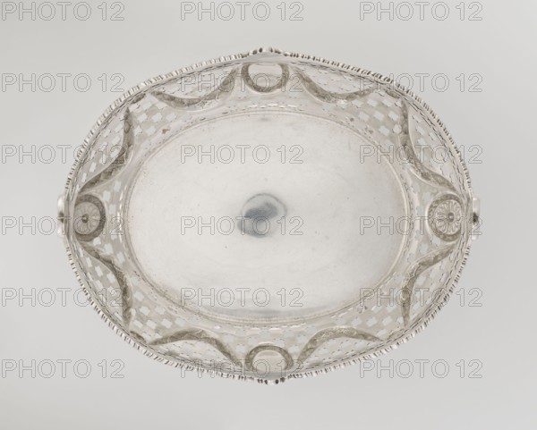 Openwork fruit basket by Harris, CS & Sons London; goldsmith's firm; fl. 1817–1933 and Charles Stuart Harris goldsmith; fl. 1852–1897; inscription/association with Ignacy Jan Paderewski 1860–1941. Oval openwork fruit basket supported on four palmette legs with the lower edge decorated with U-sections; sides feature openwork circle and trellis motifs with applied laurel garlands, medallions and rosettes; wavy-cut upper edge decorated with a godron ribbon, raised on the longer axis and decorated with acanthus leaves, on the shorter axis with palmettes. Materials: presumably silver; decorative elements include palmettes, bay leaves, garlands, godron ornaments, medallions, rosettes. Date: 1896/1897. Place: London, England, Great Britain. Function: vessel fruit basket. Dimensions: height 11.2 cm; width 23 cm; depth 18.5 cm; weight 645 g., A round metallic dish photographed from above with a central smooth shallow basin surrounded by an openwork rim featuring repeating cutout shapes and scal...