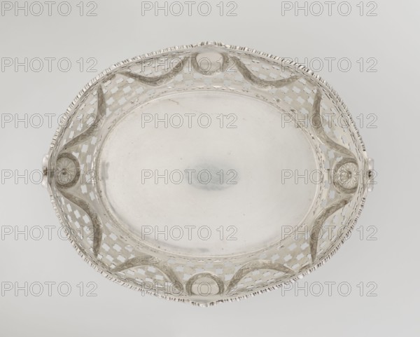 Openwork fruit basket by Harris, CS & Sons London; goldsmiths, fl. 1817–1933 and Charles Stuart Harris fl. 1852–1897. Oval openwork fruit basket supported on four palmette legs with a lower edge decorated with C-sections. Sides feature openwork composed of circle and grid motifs with applied laurel garlands, medallions, and rosettes. Upper edge cut in a wavy pattern and decorated with a godron ribbon; raised on the longer axis and decorated with acanthus leaves, and on the shorter axis with palmettes. Contains palmettes, bay leaves, garlands, godron ornaments, and medallions., A shallow round decorative dish viewed from above with an openwork rim featuring repeating quatrefoil-shaped cutouts and undulating raised bands; the central flat surface is smooth and reflective. The rim has a notched edge and two small protruding handles with floral rosette motifs where they attach to the rim. The object appears metallic with a predominantly silver color and subtle variations including slightly...