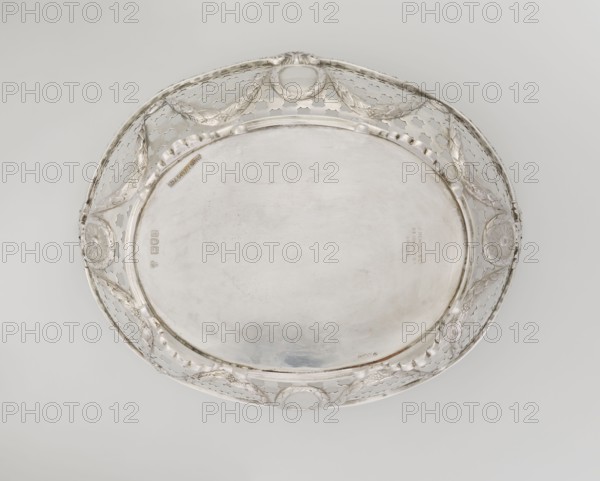 Openwork fruit basket, Harris, CS & Sons London; goldsmith's firm; fl. 1817–1933 and Charles Stuart Harris goldsmith; fl. 1852–1897. Oval openwork fruit basket supported on four palmette legs with a lower edge decorated with C-sections. Sides feature openwork circle-and-grid motifs with applied laurel garlands, medallions, and rosettes. Upper edge is cut in a wavy pattern and decorated with a godron ribbon; raised on the longer axis and decorated with acanthus leaves, and on the shorter axis with palmettes. Includes palmettes, medallion, bay leaves, garlands, and godron ornament., A round, shallow metal tray with a wide decorative rim sits against a pale background; the tray is predominantly silver in color with areas of slightly darker patina and faint discoloration, and the rim features an openwork pattern with a repeating quatrefoil-like motif and a few garland-like swags connecting oval cartouche shapes, along with raised scalloped edging and several round floral rosettes.