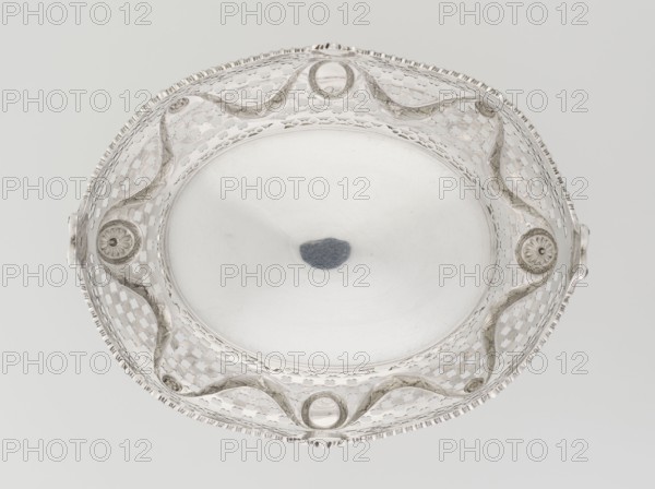 Fruit basket by Harris, CS & Sons London; goldsmith's firm, fl. 1817–1933 and Harris, Charles Stuart goldsmith, fl. 1852–1897; associated with Ignacy Jan Paderewski 1860–1941. Oval openwork fruit basket supported on four palmette legs with lower edge decorated with U-sections; openwork sides composed of circle and trellis motifs with applied laurel garlands, medallions and rosettes; wavy-cut upper edge decorated with a godron ribbon; raised on the longer axis and decorated with acanthus volutes, on the shorter axis with acanthus leaves. Materials/technique: openwork metal goldsmithing. Date: 1896–1897. Place: London, England, Great Britain. Classification: vessel. Dimensions: height 15 cm; width 29.2 cm; depth 23.3 cm; weight 1075 g., An overhead view of an ornate shallow bowl with a circular central field surrounded by a pierced decorative border; the bowl appears metallic with a silvery sheen and shows a small dark bluish-gray irregular reflection near the center; the border features...