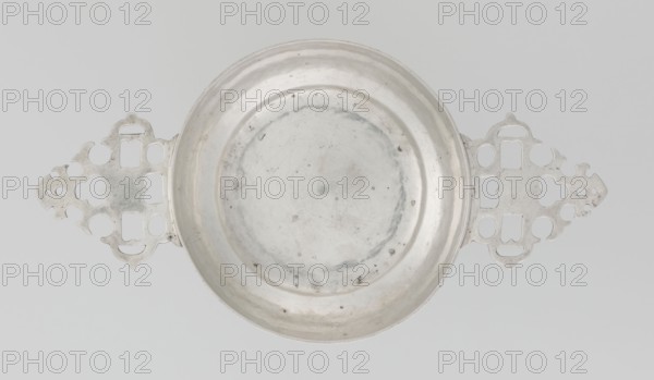 Porridge bowl porringer, maker: Vander, CJ, Ltd goldsmith's firm; established 1856; makers: Vander, Henry & Vander Arthur goldsmiths. Function: porringer/porridge dish. Description: hemispherical silver bowl with flat bottom; two flat triangular openwork handles with geometric decoration; geometric ornament; smooth surface; openwork. Date: 1912/1913. Place: Great Britain, England, London. Dimensions: height 4.5 cm; width 25.8 cm; depth 13.7 cm. Weight: 319 g., A shallow round metal bowl with a smooth interior surface and slight discoloration sits centered on a plain pale background, flanked on either side by ornate flat metal handles with symmetrical cutout patterns; the visible colors are silver-gray of the metal and a light gray background, with darker gray spots and marks on the bowl and subtle variations of gray on the handles.