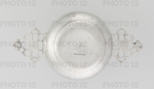 Porridge bowl porringer, maker: Vander, Henry & Vander Arthur; maker's firm: Vander, CJ, Ltd London; goldsmith's firm; active from 1856. Hemispherical silver porridge dish with flat bottom and two flat triangular openwork handles with geometric decoration; smooth surface; openwork, geometric ornament. Date: 1912–1913. Place: London, England, Great Britain. Dimensions: height 4.5 cm; width 25.8 cm; depth 13.7 cm; weight 319 g. Function: porringer porridge dish., A metallic, circular bowl-like object with a smooth convex center and a slightly raised rim sits against a light gray background, flanked on opposite sides by ornate, symmetrical filigree handles with cut-out shapes and rounded motifs; the metal surface shows scratches, scuffs, and small darker spots, and the visible colors are silver-gray and light gray.