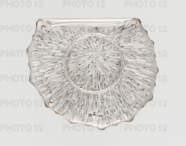 Jam or honey container stand in the shape of a stylized leaf, rolled up on one side, with wavy edges and a textured surface, supported on four legs; in the middle a basket for the container in the form of a rim for legs, made of wire imitating a twig according to the design of Christopher Dresser. Maker: Hukin & Heath Birmingham; London; goldsmiths' firm; fl. 1855–1953; makers/associates: John Thomas Heath & John Hartshorne Middleton; designer: Christopher Dresser 1834–1880. Date: 1890–1891. Place: Great Britain — England, London. Style: Aestheticism. Materials/technique: metal, wire. Function: table accessory jam or honey container stand. Dimensions: height 3.5 cm; width 18.3 cm; depth 15.5 cm; weight 267 g., A scalloped, circular object with a textured, radial pattern fills the image; the surface shows a mix of translucent and reflective areas with visible irregularities and small dark spots, and the dominant colors visible are silver, white, gray, and hints of darker gray.