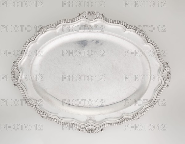 Oval Platter, Smith, Benjamin & James fl. 1809–1812 maker's role / goldsmith's firm, coats of arms, monograms, godron ornaments, shells ornament, Georgian style, acanthus ornament, George III style; oval, with smooth mirror and multi-leaf ribbed collar, edged with godron band with four shells on axes, enclosed within acanthus leaves; engraved coats of arms on collar; on the underside of the collar engraved monogram GvR, 1811/1812, Great Britain, England, London; vessel, height 3.5 cm, width 58.5 cm, depth 45.4 cm, weight 3550 g, A large ornate oval tray with a scalloped decorative rim and molded edge, rendered in metallic silver tones with areas of darker gray patina and subtle scratches across its central surface, showing a smooth slightly concave center and a raised patterned border; the rim features carved shell and scroll motifs at the top and bottom and matching ornamental handles on opposite sides, all appearing in silver and gray against a pale neutral background.