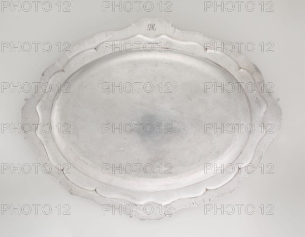Oval Platter, Smith, Benjamin & James fl. 1809–1812 maker's role / goldsmith's firm; Georgian, George III style; oval vessel with smooth mirror and multi-leaf ribbed collar edged with a godron band with four shells on axes, enclosed within acanthus leaves; engraved coats of arms on collar; underside of collar engraved monogram GvR, 1811/1812; materials and ornament: godron, acanthus, shells; inscriptions/marks: coats of arms, monogram; place: London, England; dimensions: height 3.5 cm, width 58.5 cm, depth 45.4 cm; weight 3550 g., An oval metal tray with a scalloped rim seen from above, showing a smooth central surface with visible scratches and light wear; the tray is mostly silver-gray with subtle variations in tone and slightly darker smudging near the center, and the rim has paler silver highlights and faint shadowed edges giving hints of texture.