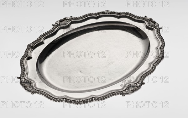 Oval platter by Benjamin & James Smith fl. 1809–1812, maker's mark/goldsmith's firm; Georgian, George III style; oval silver vessel with smooth mirror and multi-leaf ribbed collar edged with a godron band and four shells on axes, enclosed within acanthus leaves; godron ornaments, acanthus ornament; engraved coats of arms on collar; underside of collar engraved monogram GvR, dated 1811/1812; monograms, coats of arms; shells ornament; provenance noted; London, England, Great Britain; height 3.5 cm, width 58.5 cm, depth 45.4 cm, weight 3550 g., An oval metal serving tray with an ornate raised rim featuring scalloped edges and decorative scrollwork, showing a reflective silver surface with visible scratches and slight dents; the tray is predominantly silver and metallic gray with areas of darker gray where shadows and reflections appear, and it rests against a pale gray background.
