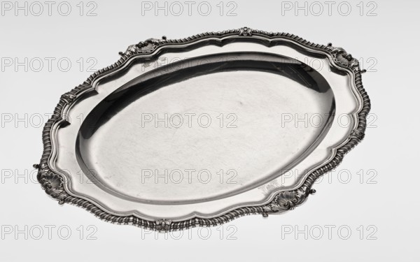 Oval platter by Benjamin & James Smith fl. 1809–1812, makers; goldsmith's firm. Georgian, George III style. Oval vessel with smooth mirror and multi-leaf ribbed collar, edged with a godron band with four shells on axes, enclosed within acanthus leaves. Engraved coats of arms on collar; underside of collar engraved monogram GvR, dated 1811/1812. Decorations: coats of arms, monograms, godron ornaments, shells, acanthus. Materials/technique: vessel silver. Dimensions: height 3.5 cm; width 58.5 cm; depth 45.4 cm; weight 3550 g. Place: London, England, Great Britain., A large oval metal tray with an ornate scalloped rim is shown against a plain light gray background; the tray appears reflective with a polished silver surface showing subtle scratches and surface marks, and the rim features decorative raised scrollwork and shell-like motifs with ribbed detailing in darker gray tones contrasted against the shiny silver; overall colors visible are silver, gray, and darker metallic gray shadows.