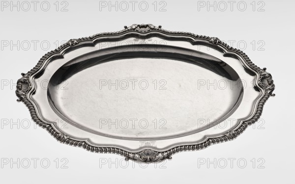 Oval Platter, Smith, Benjamin & James fl. 1809–1812 maker's role / goldsmith's firm, coats of arms, monograms, godron ornaments, shells ornament, Georgian style, acanthus ornament, George III style, oval, with smooth mirror and multi-leaf ribbed collar, edged with godron band with four shells on axes, enclosed within acanthus leaves; engraved coats of arms on collar; on the underside of the collar engraved monogram GvR, 1811/1812, Great Britain, England, London, vessel, height 3.5 cm, width 58.5 cm, depth 45.4 cm, weight 3550 g, A large oval metal tray with an ornate scalloped rim and decorative raised edging, showing a shiny reflective surface with visible scratches and fine marks; the tray appears in shades of silver and gray with darker gray reflections in the inner rim and brighter silver highlights on raised areas, and the background is a smooth very light gray.