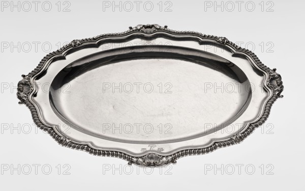 Oval platter by Benjamin & James Smith fl. 1809–1812, goldsmiths; Georgian/George III style, oval vessel with smooth mirror and multi-leaf ribbed collar edged with a godron band with four shells on axes, enclosed within acanthus leaves; godron ornaments, shells, acanthus ornament. Engraved coats of arms on collar; underside of collar engraved monogram GvR, dated 1811/1812. Materials: goldsmith's work. Dimensions: height 3.5 cm; width 58.5 cm; depth 45.4 cm; weight 3550 g. Places: Great Britain, England, London., A large oval metal tray with an ornate scalloped rim and decorative handles rests against a pale background; the tray has a reflective silvery surface with visible fine scratches and wear, and the rim features detailed embossed patterns and flowing curves in shades of silver and gray.