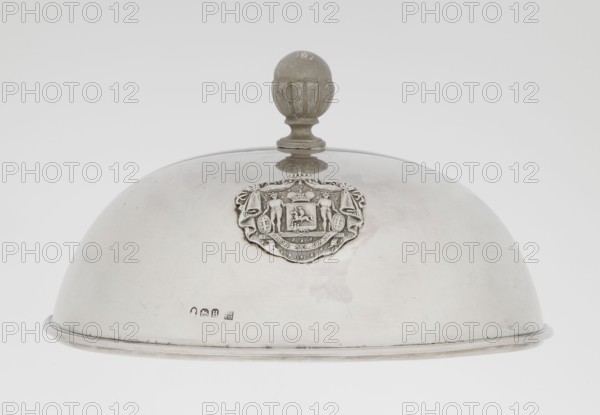 Platter lampshade, Scott, Digby & Smith, Benjamin fl. 1802–1807 goldsmith; silver hemispherical lampshade with a non-original handle, smooth with a ribbon profiled edge. On the side, a relief plaque with the Czartoryski coat of arms on a paludament, under a princely mitre, with labres and the motto Be that as it may. Handle is contemporary. 1803–1804, London, England; vessel; height 8.5 cm, diameter 15.2 cm, weight approx. 230 g., A silver dome-shaped object with a rounded finial on top, showing a decorative raised emblem at the front featuring sculpted figures and ornate detailing; the surface is metallic silver with subtle scratches and tarnish, and the finial is a slightly darker gray with textured grooves.