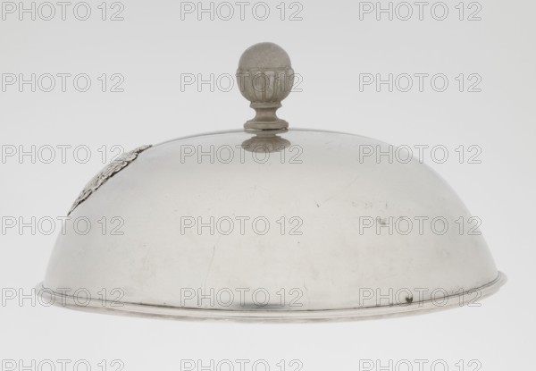 Platter lampshade, Scott, Digby & Smith, Benjamin fl. 1802–1807 goldsmith; coats of arms, Pogon Ruska coat of arms; relief decoration. Silver hemispherical lampshade with a non-original handle, smooth, with a ribbon edge, profiled. On the side, a relief plaque with the Czartoryski coat of arms on a paludament, under a princely mitre, with labres and the motto Be that as it may. The handle is contemporary. 1803/1804, London, England. Vessel; height 8.5 cm, diameter 15.2 cm, weight approx. 230 g., A curved metallic dome with a muted silvery-gray surface and a round finial on top, the finial showing a slightly darker gray and a textured, ridged design; the dome has a polished yet worn appearance with faint scratches and scuffs, a thin rim along the bottom edge, and a decorative applied element on one side that appears slightly darker silver; the background is a smooth, light gray to off-white gradient.