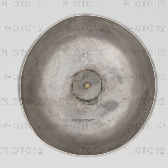 Platter lampshade, Scott, Digby & Smith, Benjamin fl. 1802–1807 goldsmith; silver hemispherical lampshade with a non-original handle, smooth, ribbon edge, profiled. Relief plaque with the Czartoryski coat of arms on a paludament, under a princely mitre, with labres and the motto Be that as it may. Handle is contemporary. 1803/1804, London, England, Great Britain. Vessel; height 8.5 cm, diameter 15.2 cm, weight approx. 230 g., A large round metallic bowl seen from above with a shallow concave interior showing a weathered, uneven surface in shades of silver and gray and a few darker gray patches, a circular metal disc with a small golden element at its center is visible in the middle, the rim is a slightly brighter silver and the background around the bowl is a pale gray.