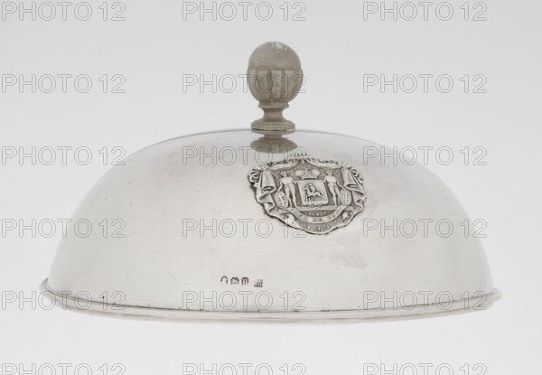 Platter lampshade by Scott, Digby & Smith, Benjamin fl. 1802–1807, goldsmith. Silver hemispherical lampshade with a non-original handle, smooth with a ribbon profiled edge. On the side, a relief plaque with the Czartoryski coat of arms on a paludament, under a princely mitre, with labres and the motto Be that as it may. The handle is contemporary. Dated 1803/1804, London, England, Great Britain. Vessel; height 8.5 cm; diameter 15.2 cm; weight approx. 230 g., A domed metal cloche with a round finial handle on top sits against a plain light background; the cloche is smooth and reflective with visible scuffs and scratches and a worn metallic sheen in silver and gray tones, and a decorative raised emblem with intricate sculpted details is attached to one side showing textured relief in similar silver-gray metal, while the finial appears slightly darker with muted gray and brownish discoloration, and the rim shows a thin darker band of metal.