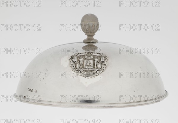 Platter lampshade, Scott, Digby & Smith, Benjamin fl. 1802–1807 goldsmith; Silver hemispherical lampshade with a non-original contemporary handle, smooth with a profiled ribbon edge. On the side, a relief plaque with the Czartoryski coat of arms on a paludamentum, under a princely mitre, with labry and the motto Badz co sie Be that as it may. 1803/1804, London, England, Great Britain. Vessel; height 8.5 cm, diameter 15.2 cm, weight approx. 230 g., A polished domed metal cover with a rounded finial on top, showing a few scuffs and small blemishes on its surface; a decorative raised emblem is affixed near the top center, featuring sculpted human figures and ornamental drapery; the finial is a duller tone and has a ribbed, rounded shape, while the dome displays reflective highlights and subtle scratches; visible colors include silver, gray, and muted beige on the finial, with a neutral white background.