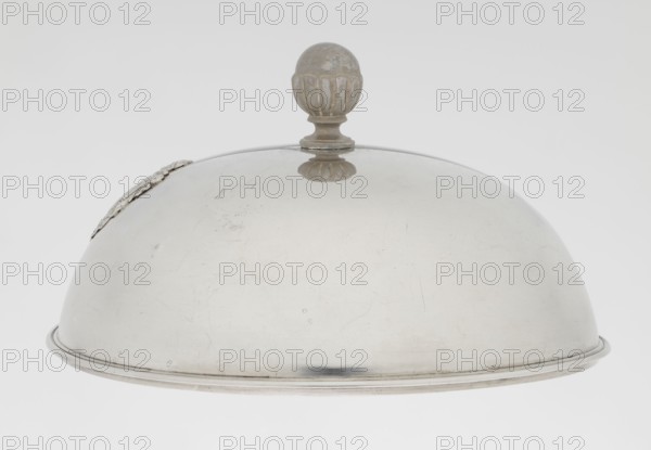 Platter lampshade, Scott, Digby & Smith, Benjamin fl. 1802–1807 goldsmith; Silver hemispherical lampshade with a non-original, contemporary handle, smooth, with a profiled ribbon edge. On the side, a relief plaque with the Czartoryski coat of arms on a paludament, under a princely mitre, with labry and the motto Badz co sie Be that as it may. 1803/1804, London, England, Great Britain. Vessel; height 8.5 cm, diameter 15.2 cm, weight approx. 230 g., A large domed metal cloche with a rounded knob handle on top; the cloche surface is smooth with visible light scratches and scuffs and a few small spots of tarnish, and there is a decorative element attached near the rim. The colors visible are silvery gray on the cloche, with areas of darker gray where the metal shows patina and shadow, and a pale off-white background.