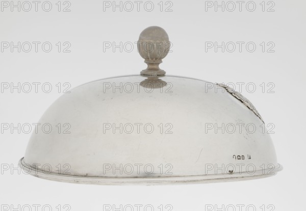 Platter lampshade by Digby & Smith, Benjamin fl. 1802–1807, goldsmith. Silver hemispherical lampshade with a non-original contemporary handle, smooth with a profiled ribbon edge. On the side, a relief plaque with the Czartoryski coat of arms on a paludament, under a princely mitre, with labres and the motto Badz co sie Be that as it may. Dated 1803/1804, London, England. Vessel; height 8.5 cm, diameter 15.2 cm, weight approx. 230 g., A polished metal dome-shaped cover with a rounded top knob, showing a smooth reflective surface with some scratches and small dark marks, a decorative element visible along one side near the rim, and a narrow raised edge around the bottom; colors visible include silver-gray of the metal, a muted darker gray on the knob and decorative element, and a light off-white background.