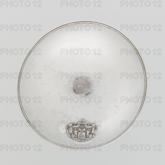 Platter lampshade by Digby & Smith, Benjamin fl. 1802–1807, goldsmith. Silver hemispherical lampshade with a non-original contemporary handle, smooth with a profiled ribbon edge. On the side, a relief plaque with the Czartoryski coat of arms on a paludament, under a princely mitre, with labres and the motto Badz co sie Be that as it may. Dated 1803–1804, London, England. Vessel. Height 8.5 cm; diameter 15.2 cm; weight approx. 230 g., A large circular metallic dish with a smooth reflective surface and a raised central boss, showing a few scratches and scuffs; near the rim at the bottom there is an ornate embossed emblem featuring decorative drapery and a small detailed scene set within a framed cartouche; the rim has a thin raised band. The visible colors are silver, light gray, and a few darker gray areas.
