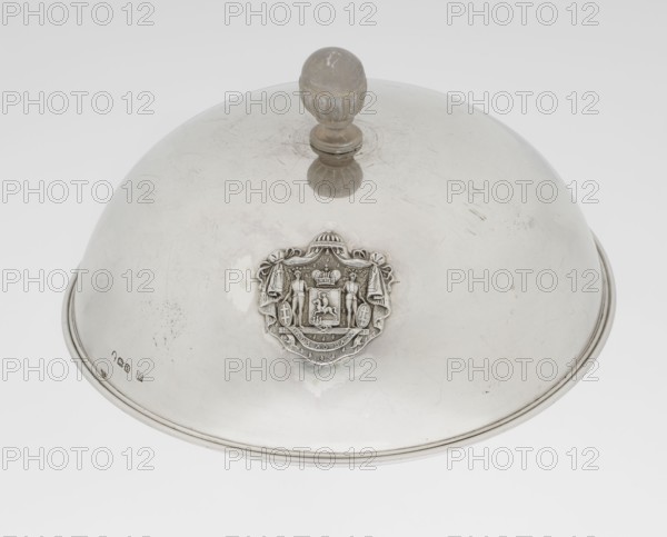 Platter lampshade, Scott, Digby & Smith, Benjamin fl. 1802–1807 goldsmith. Silver hemispherical lampshade with a non-original contemporary handle, smooth with a profiled ribbon edge. On the side, a relief plaque with the Czartoryski coat of arms on a paludament, under a princely mitre, with labres and the motto Badz co sie Be that as it may. 1803/1804. London, England, Great Britain. Vessel. Height 8.5 cm; diameter 15.2 cm; weight approx. 230 g., A domed metallic cover with a rounded knob handle on top sits against a plain light background; the cover is predominantly silver with reflective surfaces showing areas of duller gray and some small dark speckles and scratches, and features an ornate raised emblem on its side with textured details and a slightly darker silver tone, plus a thin raised rim around the base.