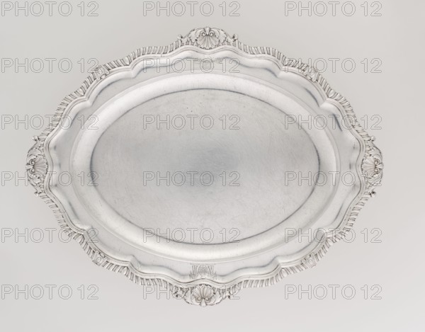 Oval platter by Benjamin & James Smith fl. 1809–1812. Silver, Georgian/George III style; oval vessel with smooth mirror and multi-leafed ribbed collar edged by a godron band with four shells on the axes, enclosed within acanthus leaves. Engraved coats of arms on the shorter axis of the collar; underside of the collar engraved monogram GvR, dated 1811/1812. Materials/technique: silver; place: London, England, Great Britain. Dimensions: height 3 cm; width 47 cm; depth 37.5 cm; weight 2420 g., A large ornate oval metal tray fills the image, showing a smooth central surface with visible fine scratches and a raised decorative rim with scalloped edges and embossed flourish patterns; the tray appears in shades of silver and gray against a pale off-white background.