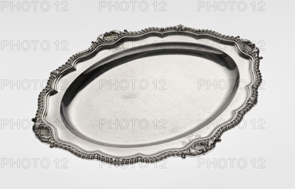 Oval platter by Benjamin & James Smith fl. 1809–1812, goldsmiths. Georgian/George III style; oval vessel with smooth mirror and multi-leafed ribbed collar, edged with a godron band with four shells on the axes, enclosed within acanthus leaves. Engraved coats of arms on the shorter axis of the collar; underside of the collar engraved monogram GvR, dated 1811/1812. Materials: silver. Place: London, England, Great Britain. Dimensions: height 3 cm; width 47 cm; depth 37.5 cm; weight 2420 g., A large oval metal tray with an ornate scalloped rim and decorative raised edges, showing a smooth interior surface with visible fine scratches and scuffs; the tray appears metallic silver with darker gray shadows along the inner rim and in recessed areas, and there are highlights of bright silver on raised decorative details and the central flat area.