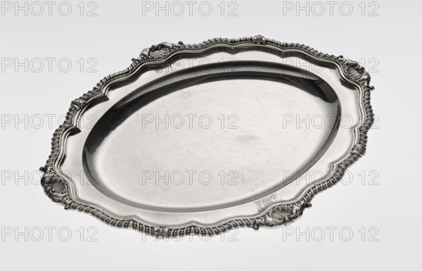 Oval platter by Benjamin & James Smith fl. 1809–1812, maker's mark; Georgian/George III style; silver vessel, oval, with smooth mirror and multi-leafed ribbed collar edged with a godron band with four shells on the axes, enclosed within acanthus leaves; engraved coats of arms on the shorter axis of the collar; underside of the collar engraved monogram GvR, dated 1811/1812; ornament: shells, godrons, acanthus leaves; dimensions: height 3 cm, width 47 cm, depth 37.5 cm; weight 2420 g; London, England, Great Britain., A large oval metal platter with an ornate raised rim featuring shell-like and rope-like decorative motifs, visible scrollwork, and embossed floral accents; the central surface is smooth with visible fine scratches and a slightly reflective finish. The colors visible are silver, gray, and hints of darker gray in the shadows and detailing.