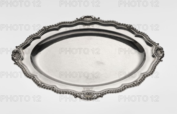 Oval platter by Benjamin & James Smith fl. 1809–1812, maker; Georgian/George III style. Silver vessel, oval with smooth mirror and multi-leafed ribbed collar edged with a godron band with four shells on the axes, enclosed within acanthus leaves. Collar engraved with coats of arms on the shorter axis; underside of the collar engraved monogram GvR, dated 1811/1812. Ornaments: coats of arms, monograms, shells, godron ornaments, acanthus leaves. London, England, Great Britain. Dimensions: height 3 cm; width 47 cm; depth 37.5 cm; weight 2420 g., A decorative oval metal tray with an ornate raised rim featuring shell and rope-like motifs, a scalloped edge, and embossed decorative flourishes at the sides and ends; the tray surface is smooth but shows visible fine scratches and scuffs; the object appears reflective and metallic, showing shades of silver and gray against a pale neutral background.