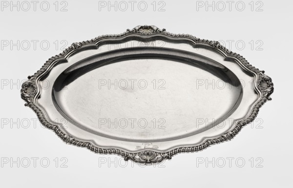 Oval platter by Benjamin & James Smith fl. 1809–1812, maker's mark; Georgian/George III style; silver; oval with smooth mirror and multi-leafed ribbed collar edged with a godron band and four shells on the axes, enclosed within acanthus leaves; engraved coats of arms on the shorter axis of the collar; underside of the collar engraved monogram GvR, dated 1811/1812; engraved shells, godron ornaments, acanthus leaves; vessel; height 3 cm, width 47 cm, depth 37.5 cm, weight 2420 g., A large ornate oval metal tray with a polished surface, showing a few fine scratches and scuffs, surrounded by an intricately molded rim with a scalloped edge and decorative shell- and scroll-like motifs at intervals; the tray appears reflective and metallic silver in color, with darker silver shadows in the recessed areas and along the inner rim, and lighter silver highlights on the raised decorative elements.